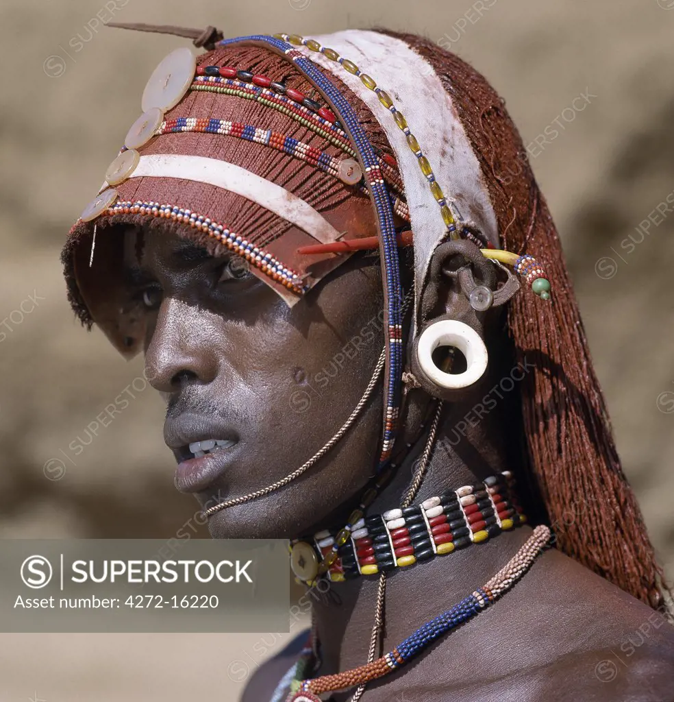 A Samburu warrior resplendent with long, braided, Ochred hair.  The braids at the front have been fashioned in a protruding fringe rather like a sunshade. The cloth on top keeps the braids in place. The round ear ornaments of the warriors are made of ivory.  Samburu warriors are vain and proud, taking great trouble over their appearance.