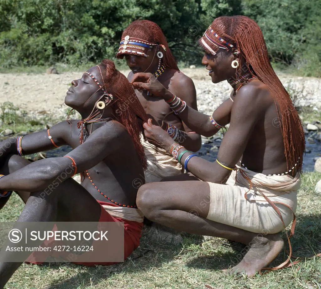 Two Samburu warriors dress the braids of a colleague. Long braids of Ochred hair distinguish warriors from other members of their society. The warriors are vain and proud, taking great trouble over their appearance.  Round ornaments, often made of ivory, adorn the pierced and extended earlobes of warriors.