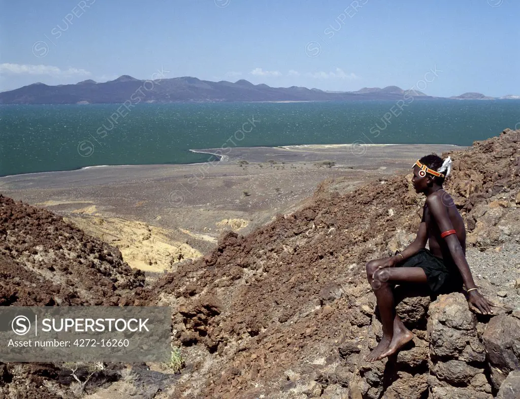 A young Turkana man looks out over Lake Turkana, often referred to as the Jade Sea due to the colour of its alkaline water.  The barren, windswept country at this southeast corner of the lake is strewn with basalt lava boulders. 160 miles long and between 8 and 27 miles wide, Lake Turkana is one of the oldest and largest lakes of Africas Great Rift Valley system.