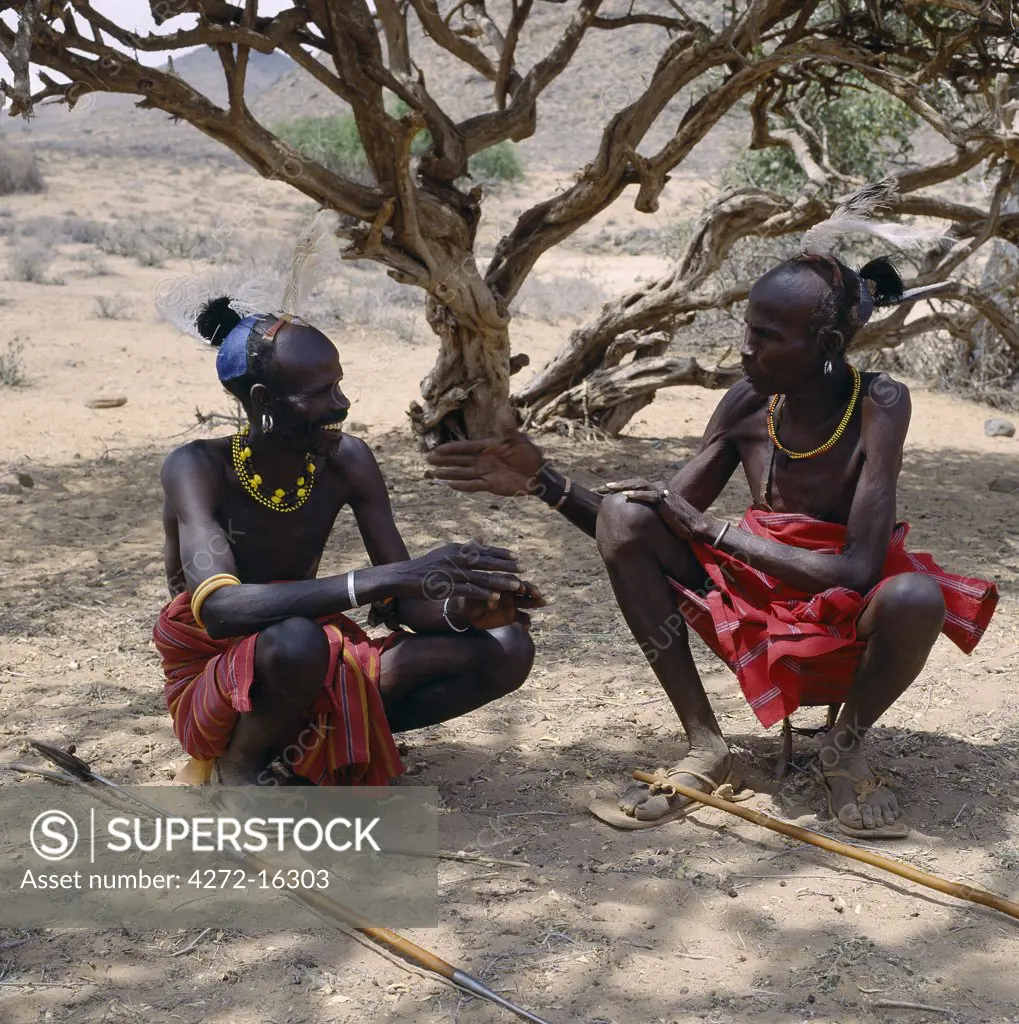 Two Turkana men in traditional attire relax in the heat of the day under a shady tree.  Every man will have a wooden stool, which doubles up as a pillow at night to protect his clay hairdo. Men will never sit on the ground; only women and children are permitted to do so.