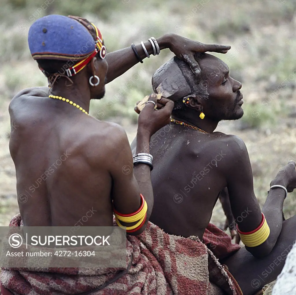 An expert hairstylist smears clay on the crown of a man's head, then fashions it into an elliptical bun before colouring it with natural  pigments.  The elliptical clay bun, so characteristic of the Turkana, is now dying out.