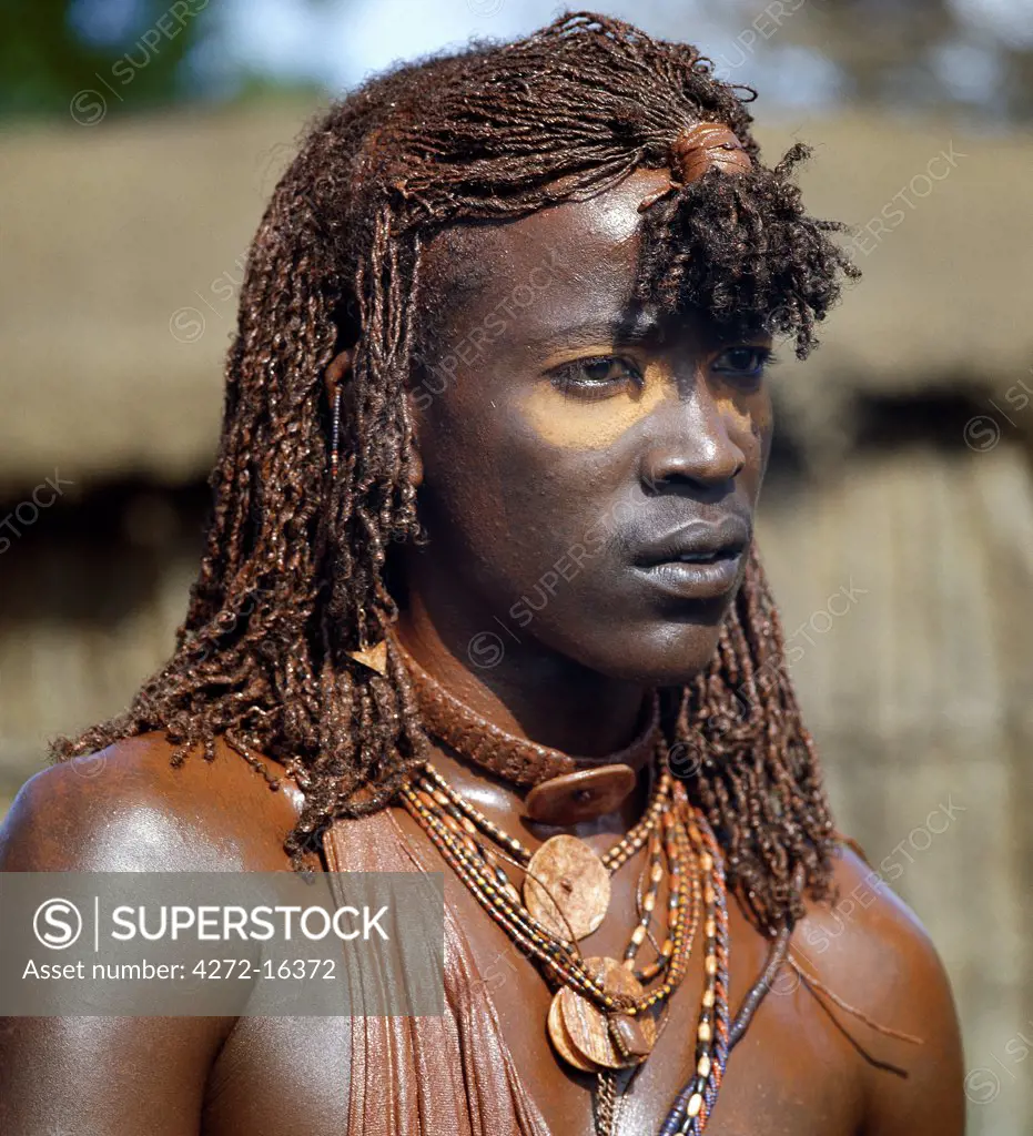 A Maasai warrior with his long braids and body coated with red ochre mixed with animal fat. He has put ochre dust round his eyes to enhance his appearance ready for a dance.  The singular hairstyles of Maasai warriors sets them apart from other members of their society.