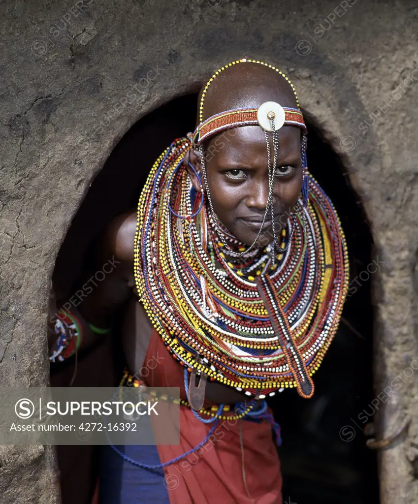 A young Maasai girl in all her finery pauses at the entrance to her mother's home. The wall and roof of the house are plastered with a mixture of cow dung and soil.