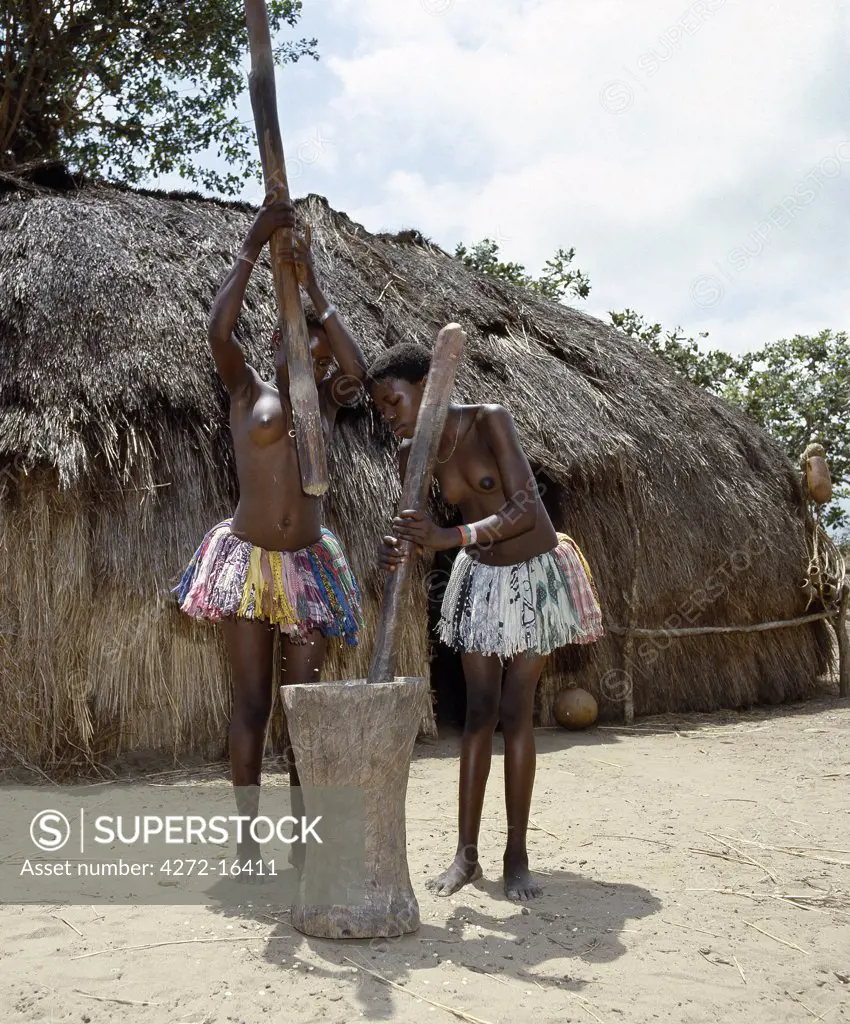 Two Giriama girls pound corn outside their home using a large wooden mortar and pestles.  Their small skirts are made from strips of printed cotton material - a traditional dress of Giriama women and children.