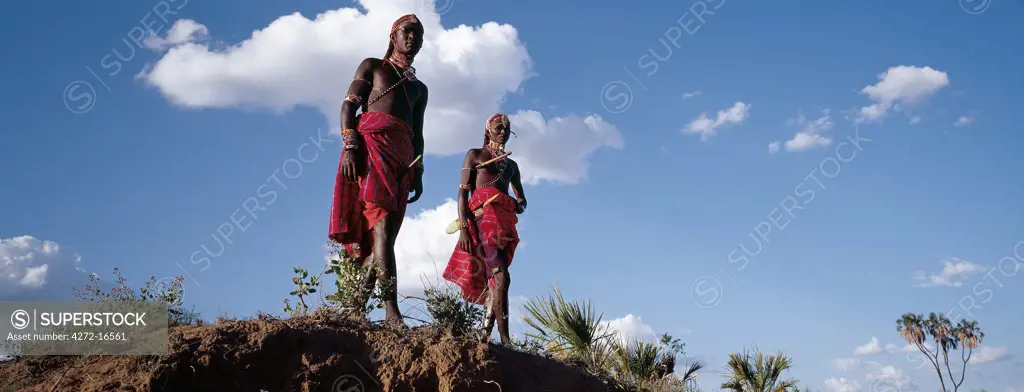Two Samburu warriors resplendent with long Ochred braids and beaded ornaments relax in typical pose beside a river bank.