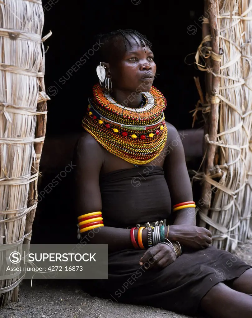 A Turkana woman, typically wearing many layers of bead necklaces and a series of hooped earrings with an pair of leaf-shaped earrrings at the front, sits in the entrance to her hut.