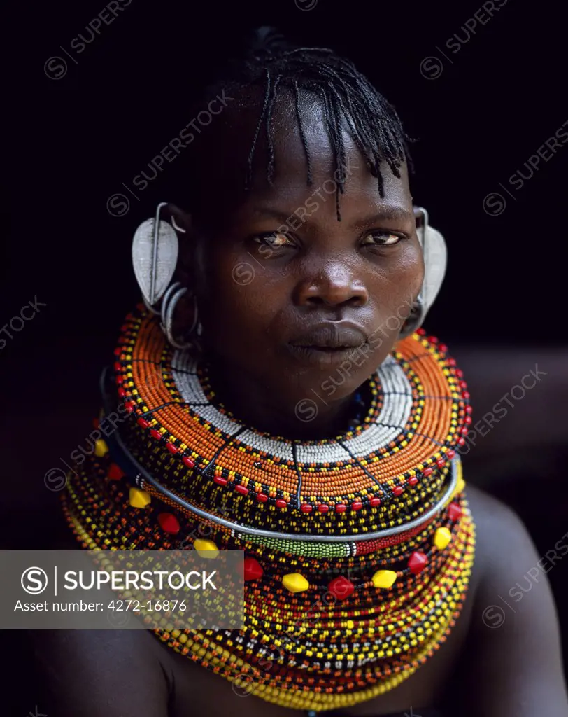 A Turkana woman sitting in the doorway of her hut.  Her heavy mporro braided necklace identifies her as a married woman.  Typical of her tribe, she wears many layers of bead necklaces and a beaded headband.