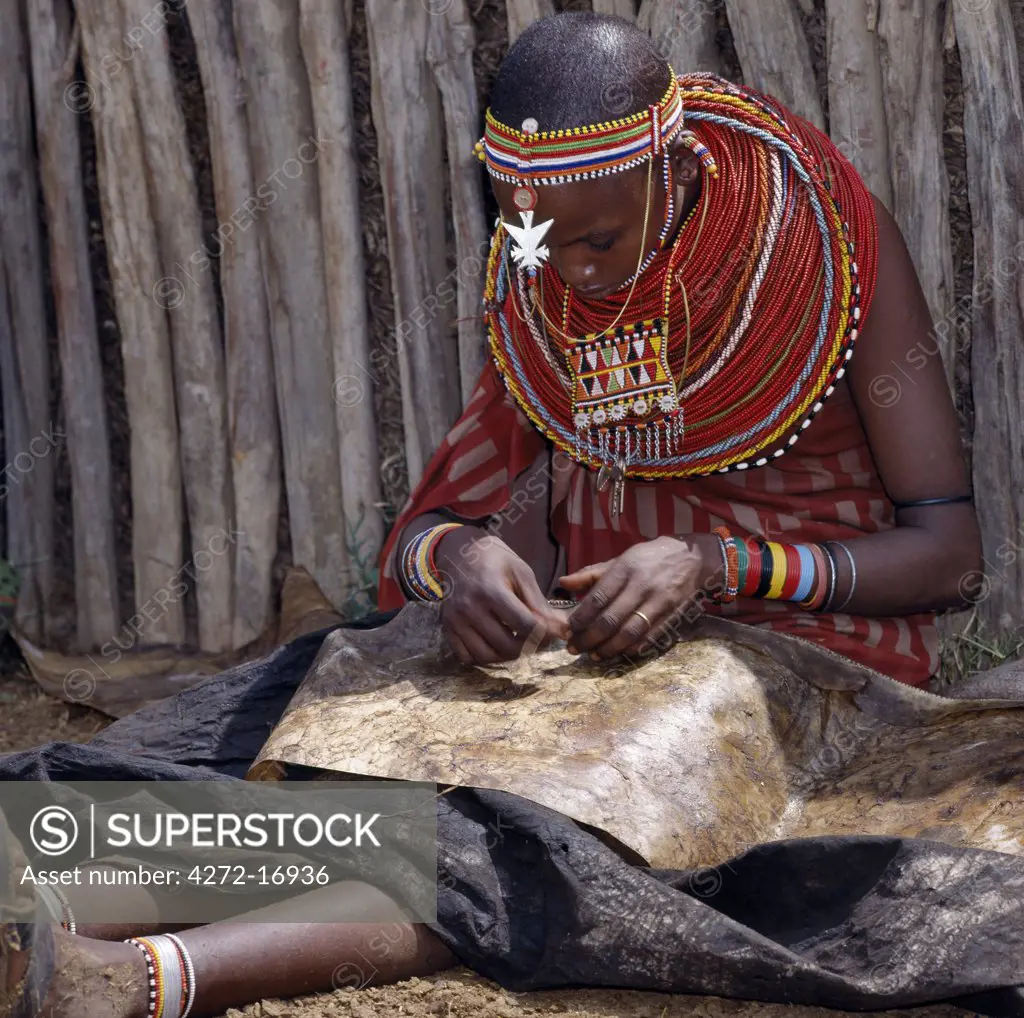 A Samburu woman sews a leather cloak for her younger brother.  For several weeks before a boy is circumcised, he must wear a charcoal-blackened cloak, which is made from three goatskins.