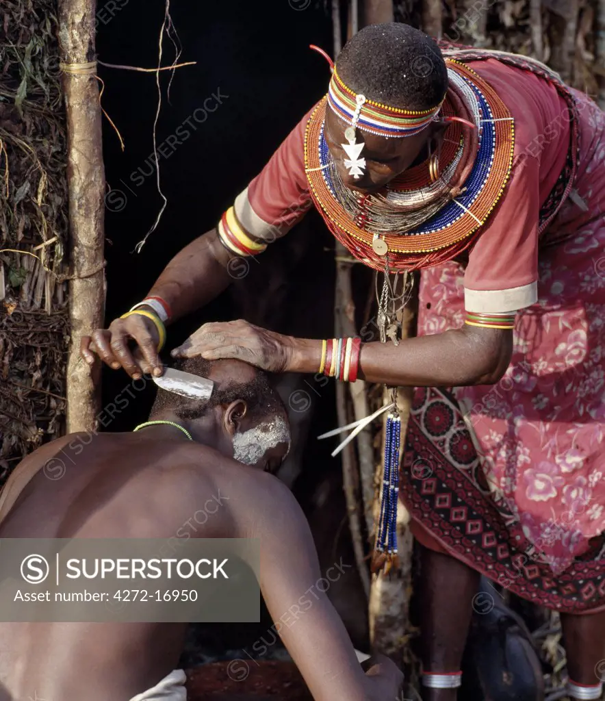 A Samburu mother shaves her sons head outside her home the day before he is circumcised. Round her neck hangs his nchipi - the distinctive decoration of every boy who participates in the circumcision ritual.