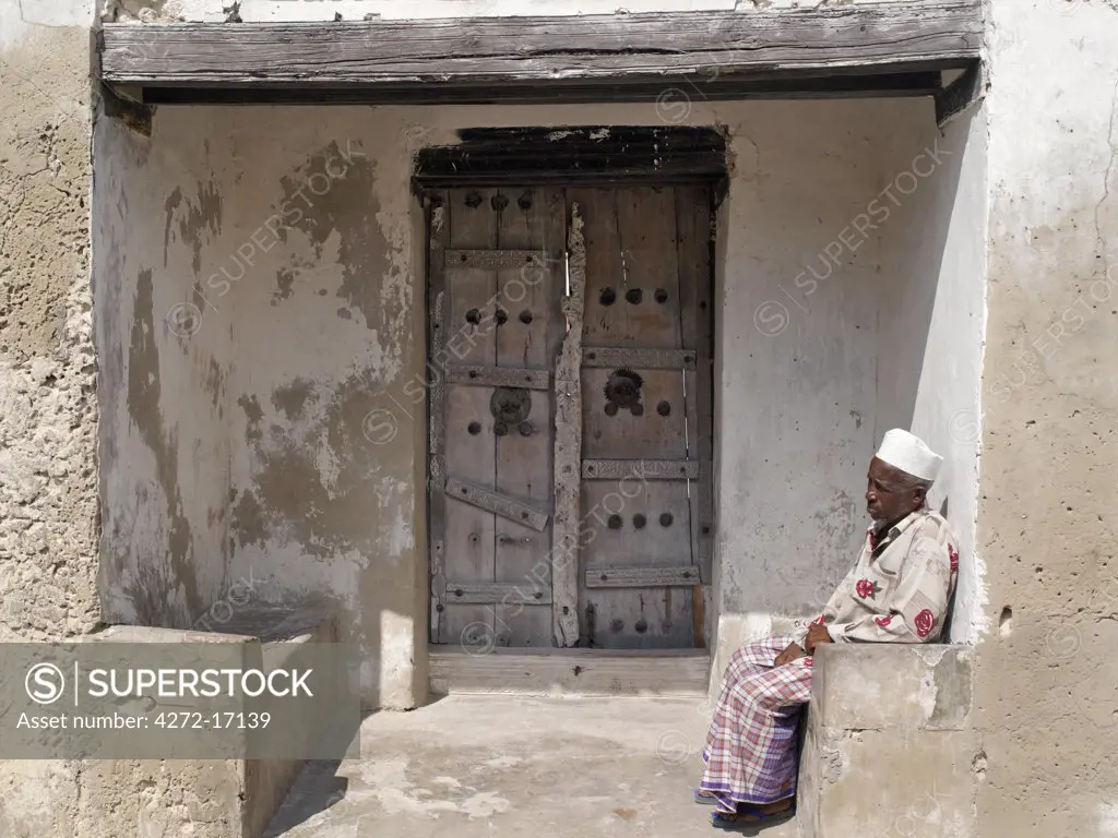 A Swahili man sits on a stone bench at the entrance to the compound of a large old house with a massive beam and decorated wooden doors.  Like most others in Faza village, the house is made of coral rag. The chequered history of Faza dates back several hundred years.