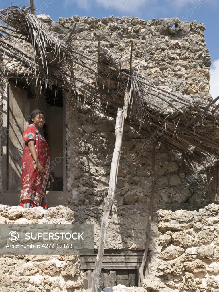 A Swahili woman in brightly coloured attire stands on the upstairs balcony of her storied house in Pate Village. All the buildings in Pate are constructed of coral rag with makuti roofs, which are a type of thatch made from coconut palm fronds. Their wooden doors and frames are often beautifully carved. Pate was established by Arabs from Arabia in the 13th century, or possibly earlier.