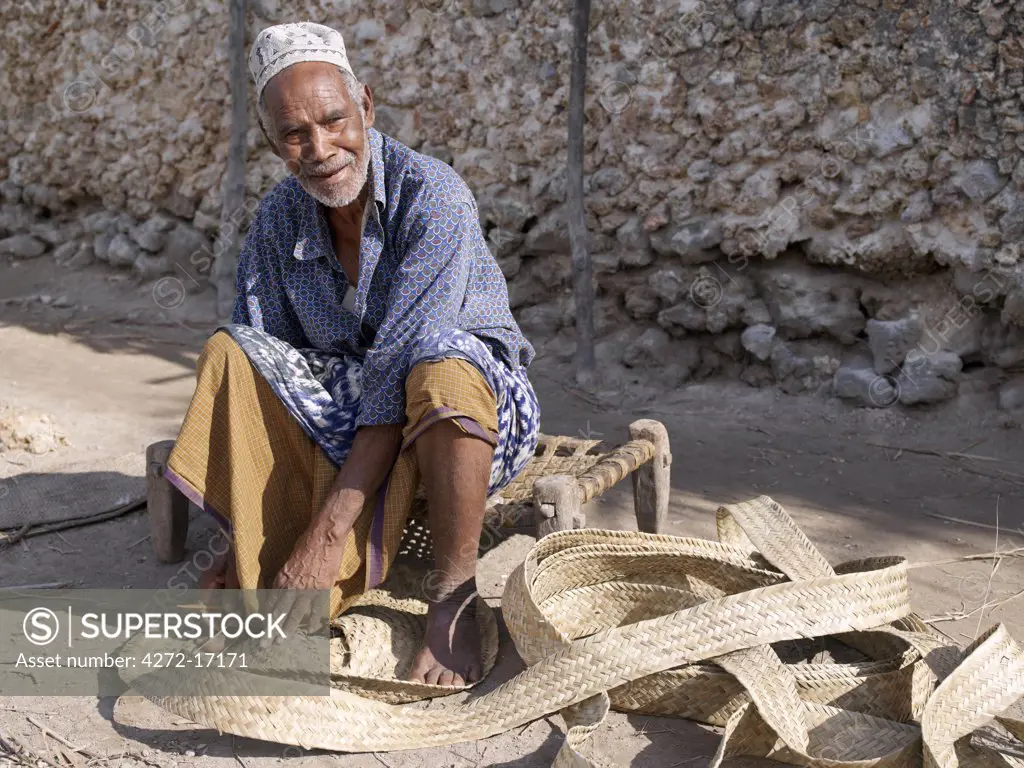Seated on a typical coast stool, a Swahili man stitches strips of woven palm fronds into a mat in one of Pate's narrow streets. All the buildings in Pate are constructed of coral rag with makuti roofs, which are a type of thatch made from coconut palm fronds.  Pate was established by Arabs from Arabia in the 13th century, or possibly earlier.