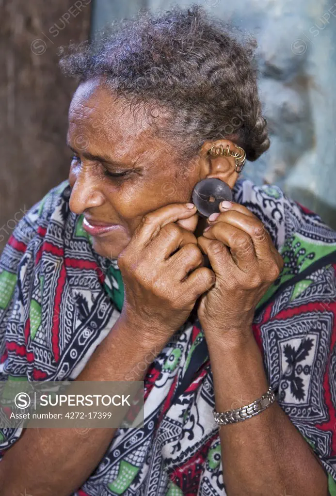Kenya, Lamu archipelago, Pate Island. An old woman on Pate Island inserts a buffalo horn earplug into her pierced earlobe. The rims of her ears are decorated with gold rings.