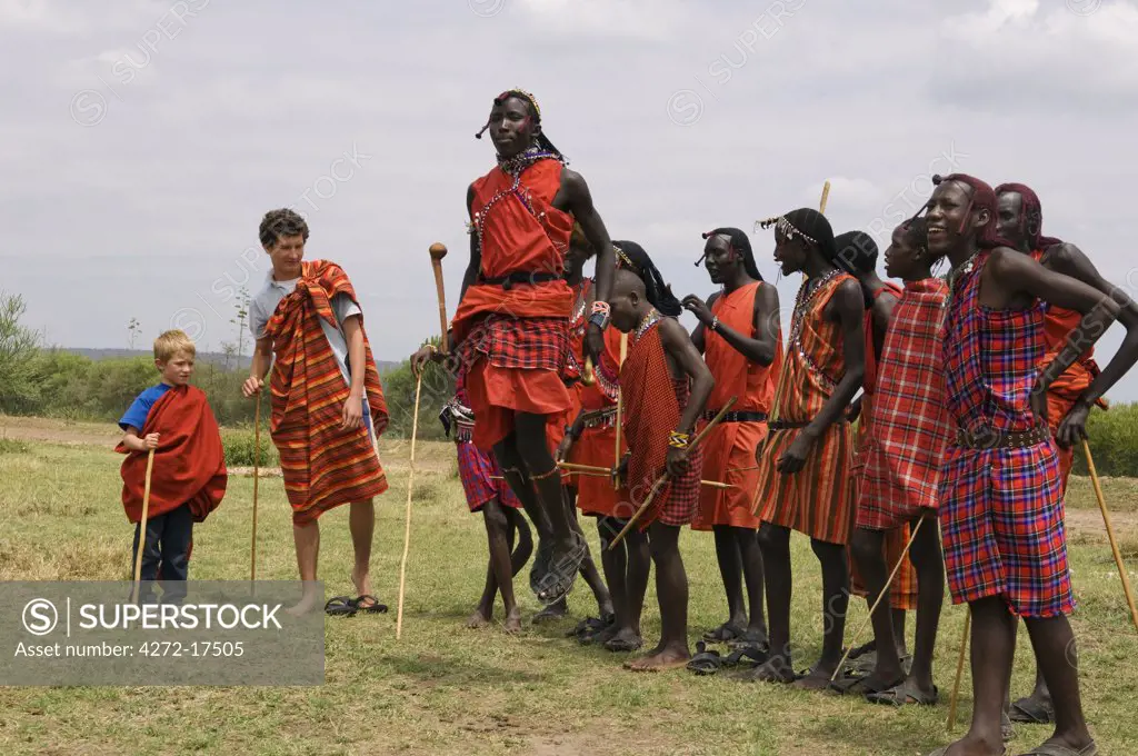 Kenya, Masai Mara National Reserve. A teenage boy on safari dances with the Maasai moran outside a manyatta at the entrance to Masai Mara Reserve.