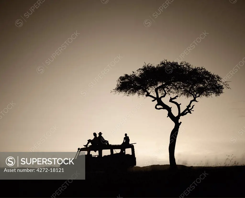 Kenya, Masai Mara Reserve.  Tourists on safari sit on top of their vehicle looking out over the savannah at dawn.