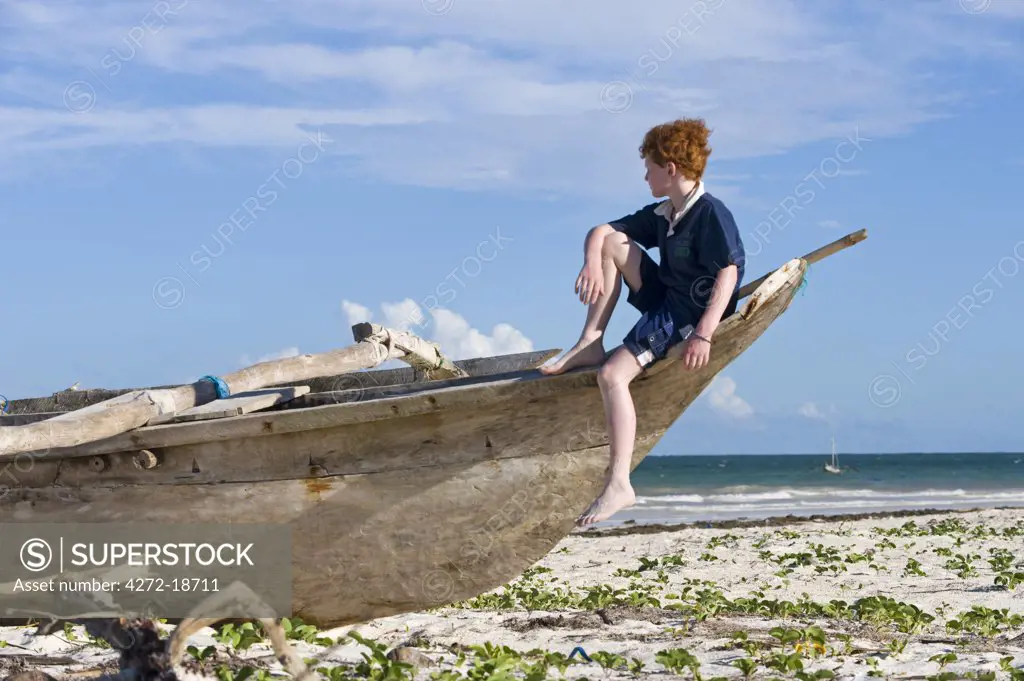 Kenya, Coast, Diani Beach.  A boy sits on a dug-out canoe whle on holiday on Galu Beach. (MR)
