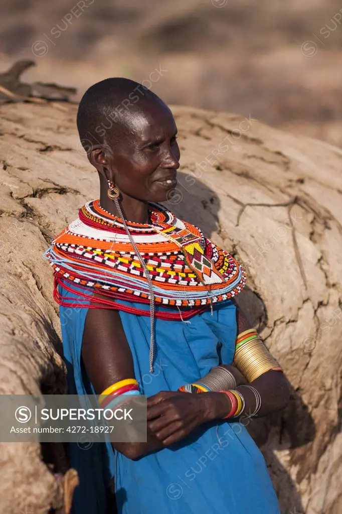 Kenya, Samburu District.  A Samburu woman, wearing intricate beaded necklaces, leans against her mud hut towards the end of the day.