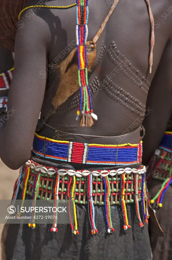 A Pokot woman in traditional attire with patterned cicatrices on her back attends an Atelo ceremony. Her broad belt is made with the metacarpus and metatarsal bones of dikdiks interspersed with beads.