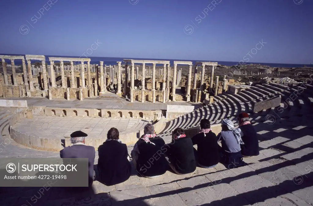 Visitors sitting on the cavea or seating area of the Theatre at Leptis Magna, showing some small temples and colonnades of cipolin columns.  Begun in the 1st Century AD and built on the site of a 3rd-5th century BC Punic necropolis, this amphitheatre was donated to the city by Annobal Rufus.  It is one of the oldest stone buildings in the ancient Roman world.