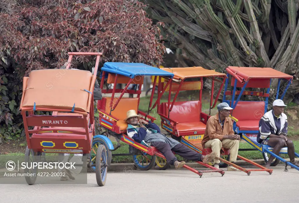 Men relax beside their brightly painted rickshaws or pousse pousse while waiting for business on the side of a busy road in Antsirabe.  It is the rickshaw or pousse pousse capital of the island. The name Pousse pousse originates from the English Push Push.