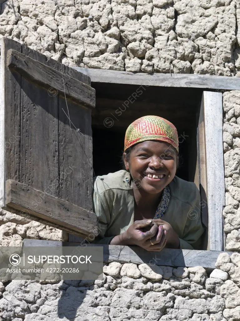 A girl wearing a Malagasy woven hat peeps out of the upstairs window of her home near Ambalavao, Madagascar