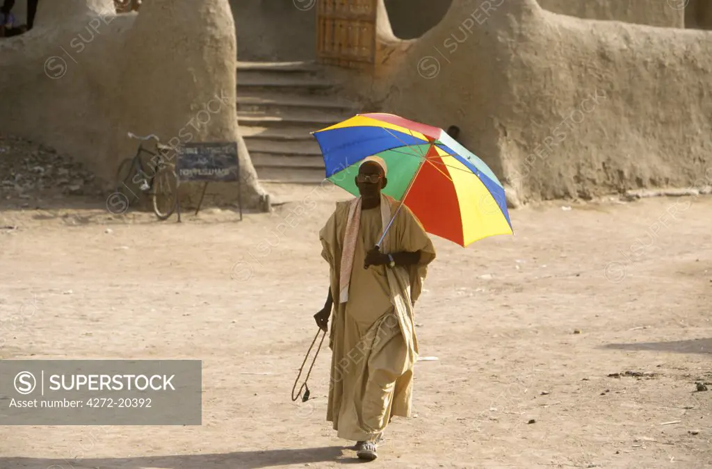 An old man uses a brightly coloured umbrella to shield himself from the Saharan sun as he walks across the main courtyard in front of the Grand Mosque
