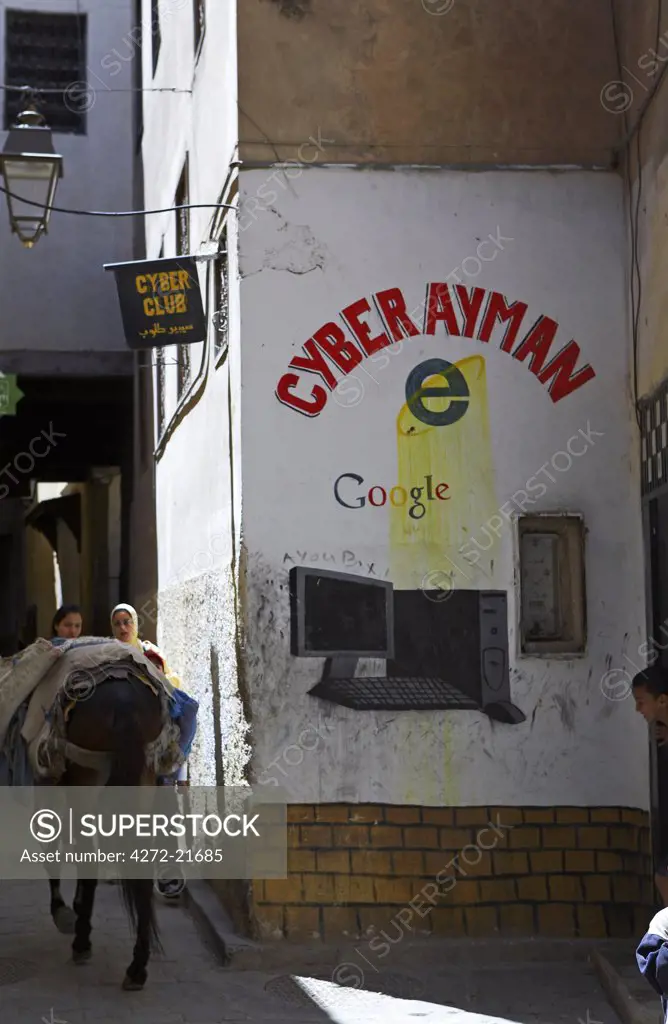 Morocco, Fes. Clash of ancient and modern as a donkey carries goods through a narrow alley, where a painted advert depicts a cyber cafe.