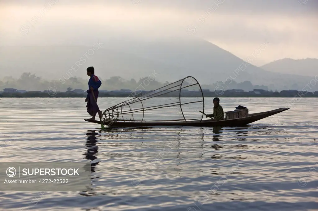 Myanmar, Burma, Lake Inle. An Intha fisherman with traditional fish trap uses an unusual leg-rowing technique to propel his flat-bottomed boat across the lake while standing.