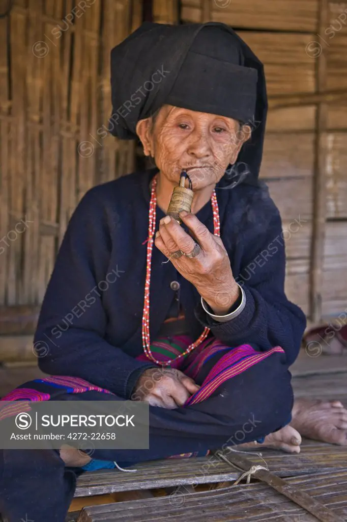 Myanmar, Burma, Wan doi. An old woman of the small Ann tribe in traditional attire sitting on the raised platform of her house as she smokes a pipe.