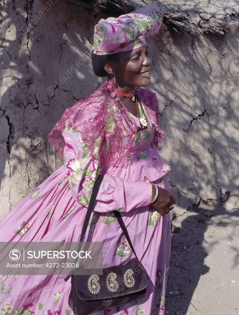 An Herero woman stands in the shade outside her home. The origins of her elaborate dress and unique hat style can be traced back to 19th century German missionaries who took exception to what they considered an immodest form of dress among the tribe.