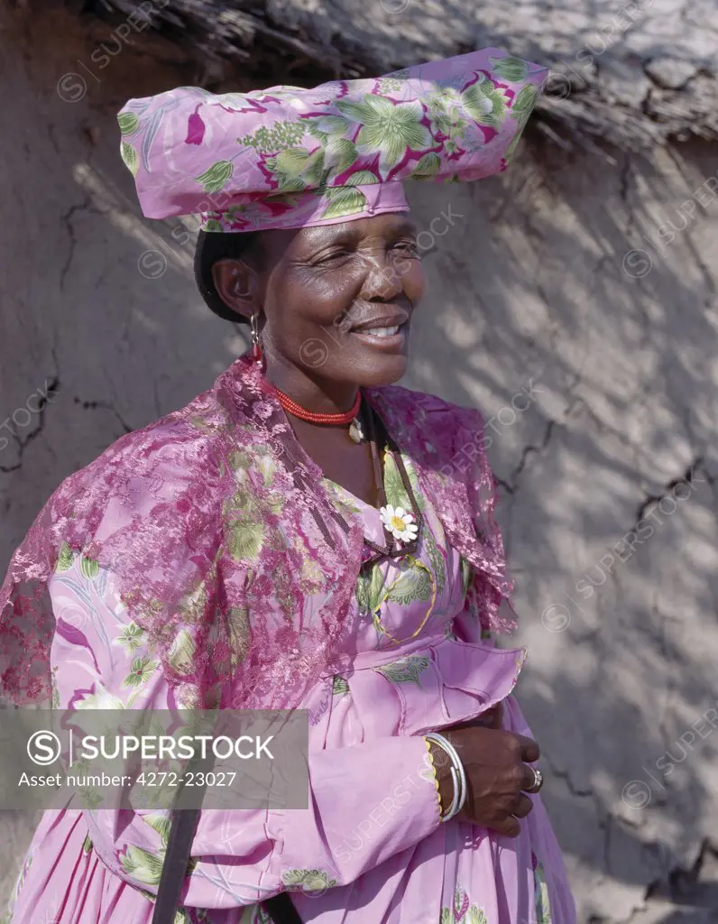 An Herero woman stands in the shade outside her home. The origins of her elaborate dress and unique hat style can be traced back to 19th century German missionaries who took exception to what they considered an immodest form of dress among the tribe.