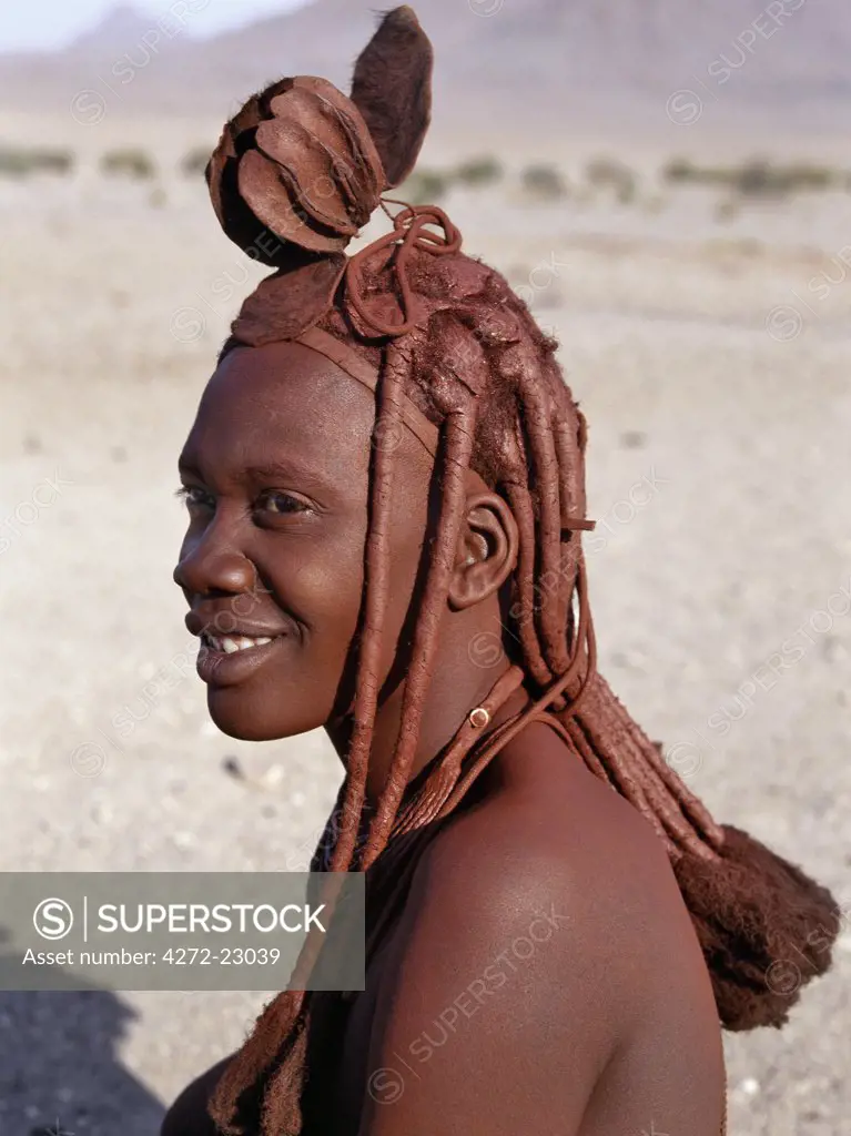 A Himba woman in traditional attire. Her body gleams from a mixture of red ochre, butterfat and herbs.  Her long hair is styled in the traditional Himba way and is crowned with a headdress made of lambskin, called erembe. The Himba are Herero speaking Bantu nomads who live in the harsh, dry but starkly beautiful landscape of remote northwest Namibia.