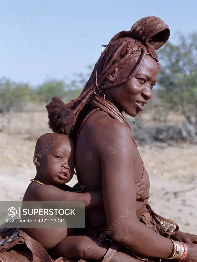 A Himba mother and child ride home on a donkey.  Their bodies gleam from a mixture of red ochre, butterfat and herbs. The womans long hair is styled in the traditional Himba way and is crowned with a headdress made of lambskin, called erembe.  The Himba are Herero speaking Bantu nomads who live in the harsh, dry but starkly beautiful landscape of remote northwest Namibia.