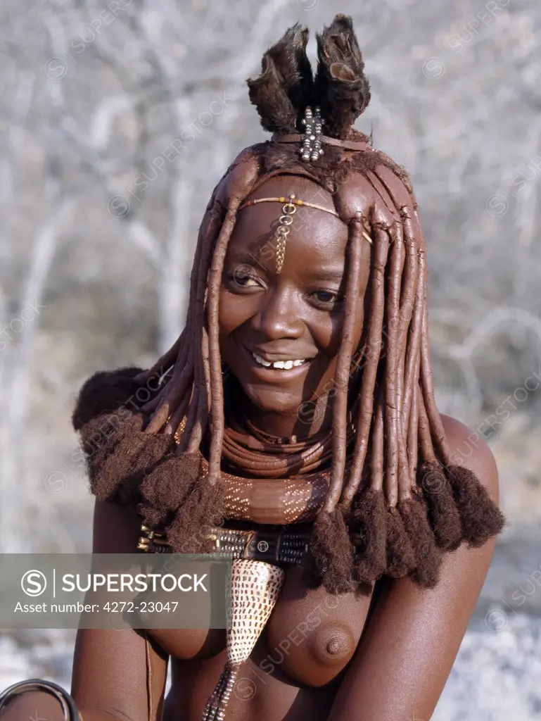 A young Himba woman in traditional attire.  Her body gleams from a mixture of red ochre, butterfat and herbs. Her long hair is styled in the traditional Himba way and is crowned with a headdress made of lambskin, called erembe.The Himba are Herero speaking Bantu nomads who live in the harsh, dry but starkly beautiful landscape of remote northwest Namibia.