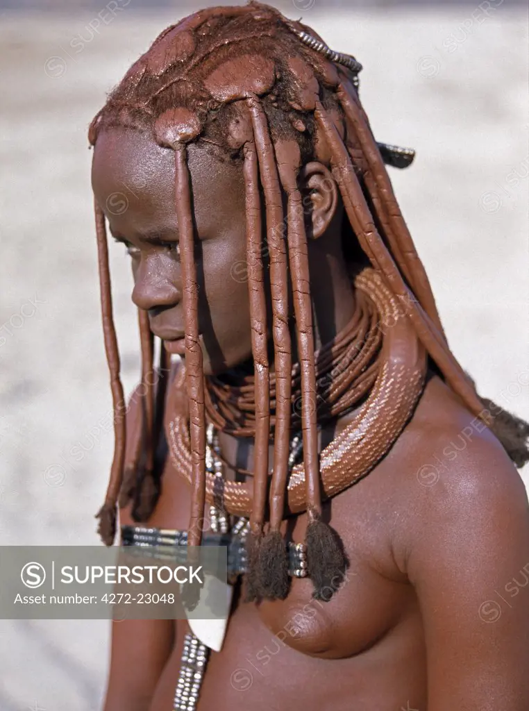 A young Himba girl in traditional attire.  Her body gleams from a mixture of red ochre, butterfat and herbs. Her long hair is styled in the traditional Himba way. The Himba are Herero speaking Bantu nomads who live in the harsh, dry but starkly beautiful landscape of remote northwest Namibia.