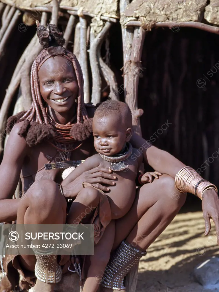 A Himba mother and baby son relax outside their dome shaped home.  Their bodies gleam from a mixture of red ochre, butterfat and herbs. The mothers long hair is styled in the traditional Himba way and is crowned with a headdress made of lambskin, called erembe.   Most married women wear a large conch shell, ohumba, between their breasts.  These prized possessions are traded from Angola and are passed down from mother to daughter.