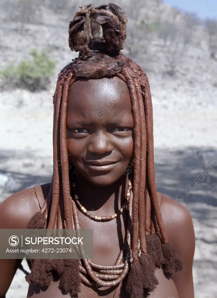 A young Himba girl in traditional attire.  Her body gleams from a mixture of red ochre, butterfat and herbs. Her hair is styled in the traditional Himba way and is crowned with a headdress made of lambskin, called erembe.