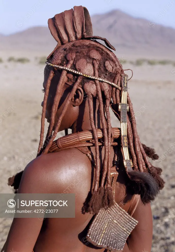 A Himba woman in traditional attire. Her body gleams from a mixture of red ochre, butterfat and herbs.  Her long hair is styled in the traditional Himba way and is crowned with a headdress made of lambskin, called erembe.