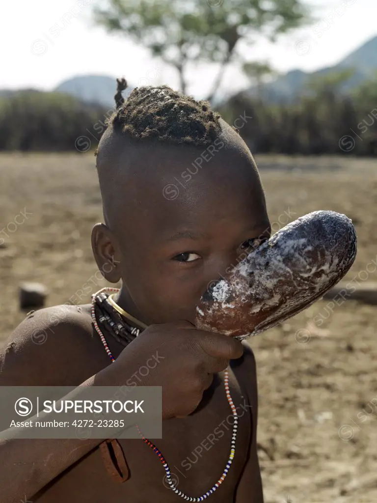 A Himba girl drinks milk from a wooden spoon. Her large, round necklace, studded with white beads, is called ombwari and is worn by both sexes.  The Himba are Herero-speaking Bantu nomads who live in the harsh, dry but starkly beautiful landscape of remote northwest Namibia.