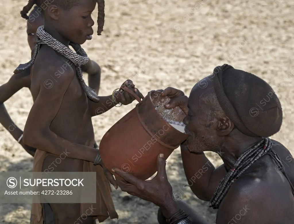 An old Himba man drinks milk from a wooden container held by one of his daughters.  The Himba are Herero-speaking Bantu nomads who live in the harsh, dry but starkly beautiful landscape of remote northwest Namibia.