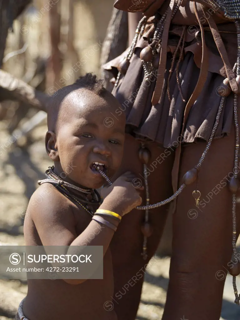 A child sucks a bead hanging from his mothers belt. A womans body is smeared with a mixture of red ochre, butterfat and herbs. The Himba are Herero speaking Bantu nomads who live in the harsh, dry but starkly beautiful landscape of remote northwest Namibia.