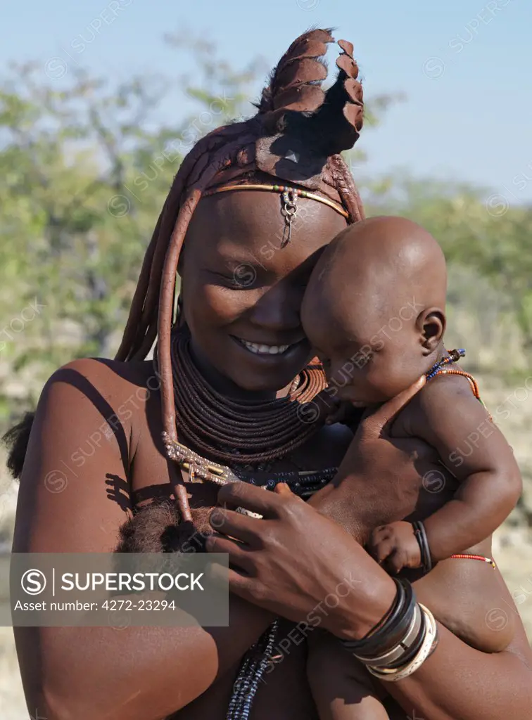 A Himba mother and child. Their bodies gleam from a mixture of red ochre, butterfat and herbs. The woman's long hair is styled in the traditional Himba way and is crowned with a headdress made of lambskin, called erembe.  The Himba are Herero-speaking Bantu nomads who live in the harsh, dry but starkly beautiful landscape of remote northwest Namibia.