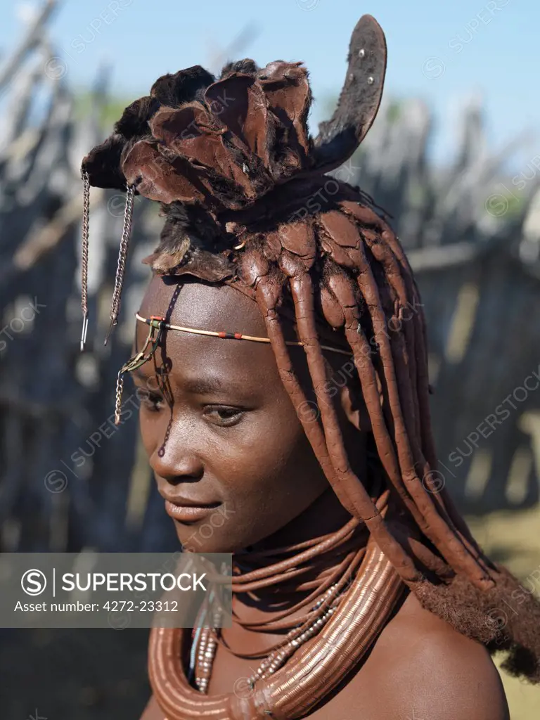 A Himba woman in traditional attire. Her body gleams from a mixture of red ochre, butterfat and herbs. Her long hair is styled in the traditional Himba way and is crowned with a headdress made of lambskin, called erembe. Her large, round white beaded necklace, called ombwari, is worn by both sexes.  Leather garments continue to be worn.