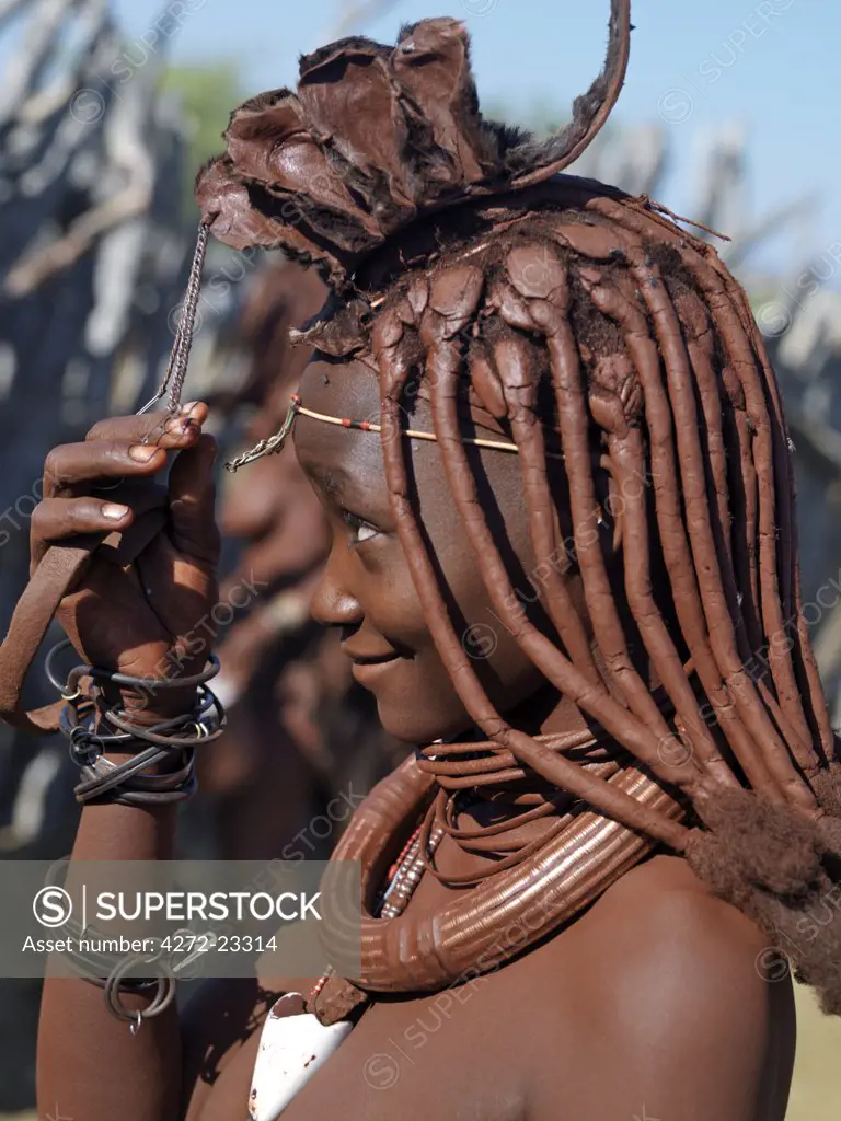 A Himba woman in traditional attire adjusts her headdress. Her body gleams from a mixture of red ochre, butterfat and herbs. Her long hair is styled in the traditional Himba way and is crowned with a headdress made of lambskin, called erembe.