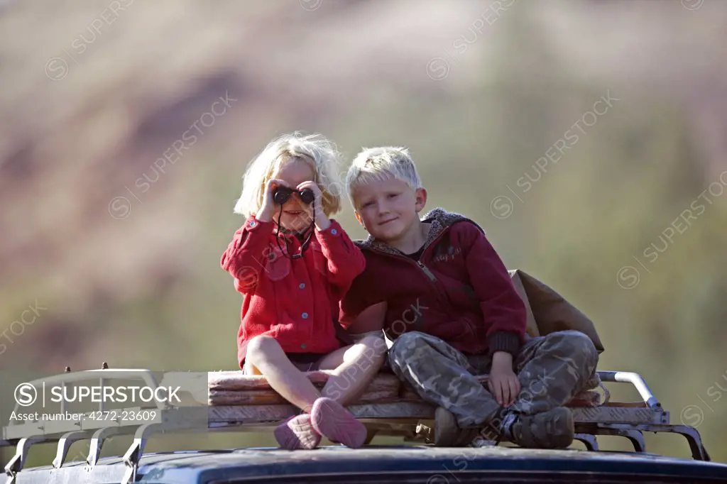 Namibia, Damaraland, Brandberg. Two young children sitting on the roof rack of a four wheel drive go game viewing down the Huab River Valley searching for elusive desert elephants with a pair of binoculars.(MR)