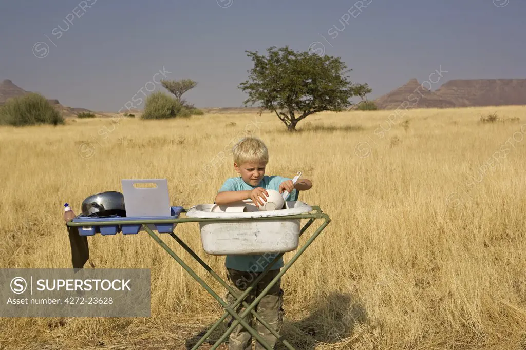Namibia, Damaraland, Etosha Region.  In the middle of the savannah a young boy washes the morning dishes on a safari dishwashing platform with grassland and acacia trees in the background. (MR)