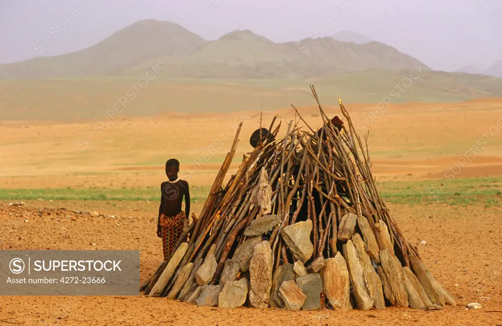 Namibia, Kaokoveld, Kaokoland. A young Himba shepherd stands beside his shelter near the Hartmann Mountains.