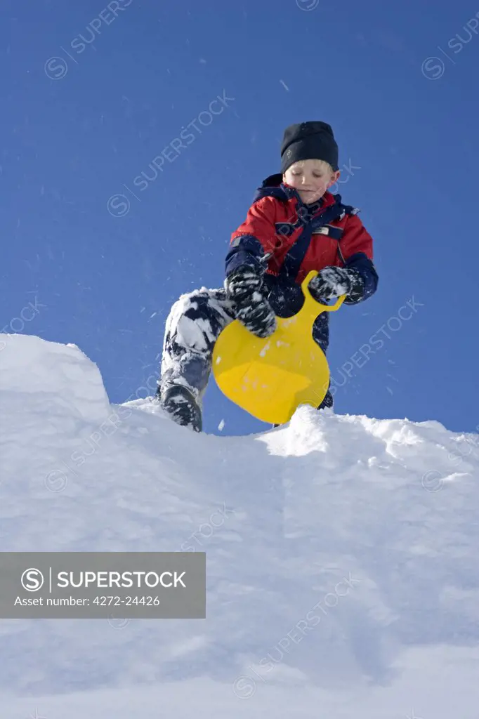 Norway, Tromso, Lyngen Alps. Young boy sits on a tobogganing seat to slide down a steep snow bank in bright winter sunshine (MR)