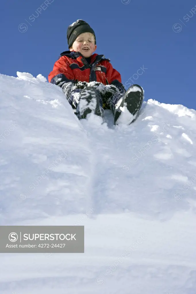 Norway, Tromso, Lyngen Alps. Young boy sits on a tobogganing seat to slide down a steep snow bank in bright winter sunshine (MR)
