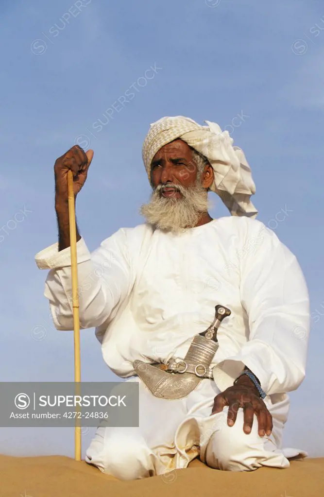 A Bedouin man kneels on top of a sand dune in the desert .  He wears the traditional Omani long white cloak or dish dash, a turban,  a ceremonial curved dagger (khanjar) and holds a short camel stick